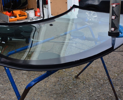 Application d'adhésif de pare-brise neuf, tenu par des ventouses bleues, près de l'arrière d'une camionnette de travail avec des outils.